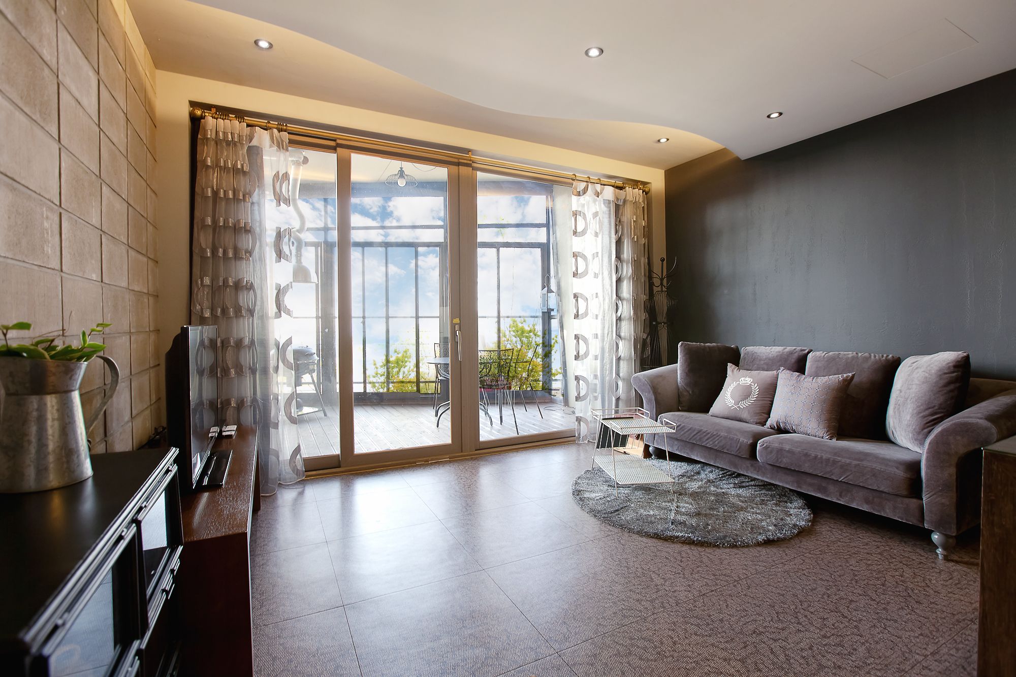 A modern living room with a grey sofa, a coffee table, and a TV on a wooden cabinet. There is a large sliding glass door leading to an outdoor balcony with a view of the sky and trees.