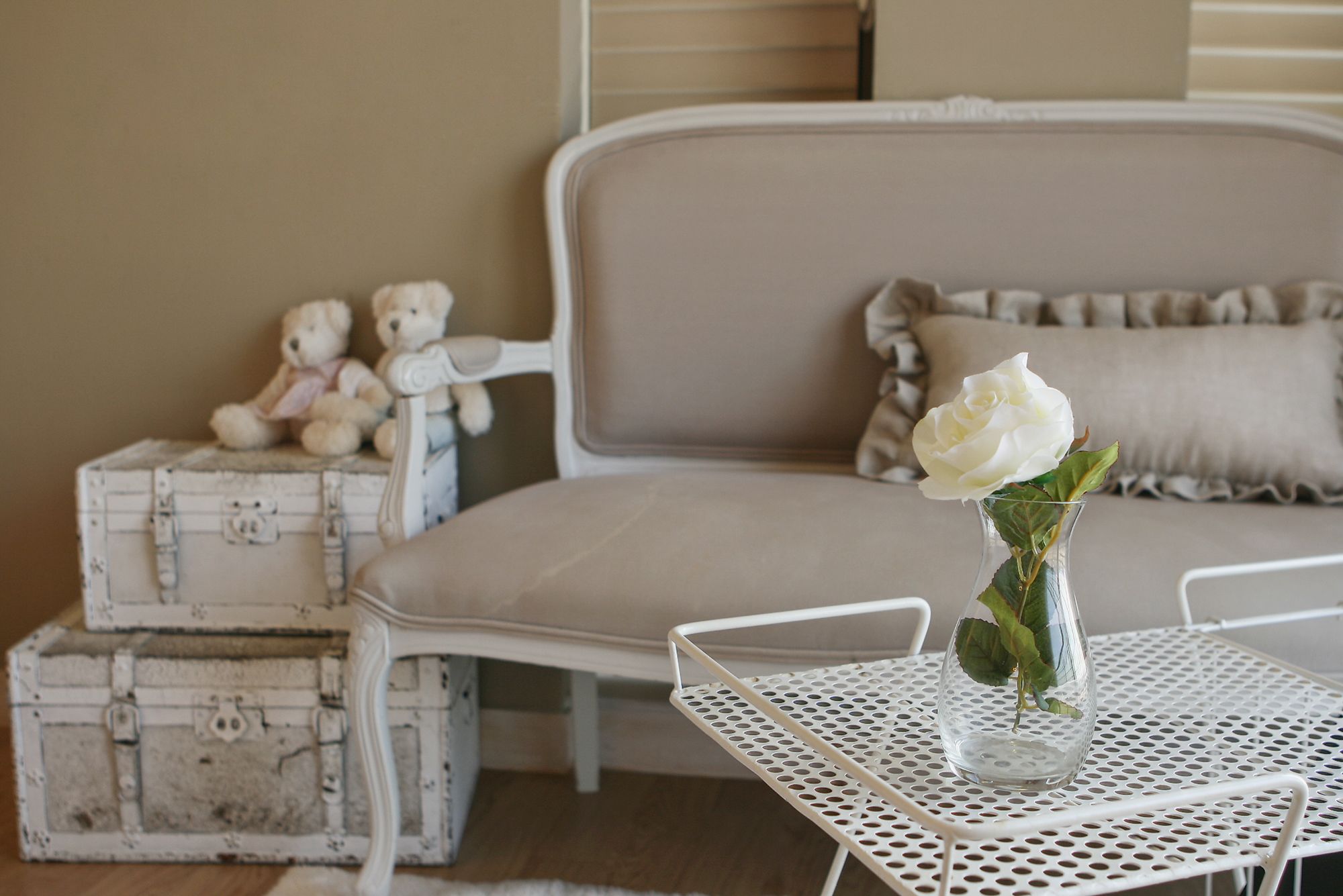 A cozy room with a bed, a small table with a vase of a white rose, and two teddy bears on a chest of drawers.