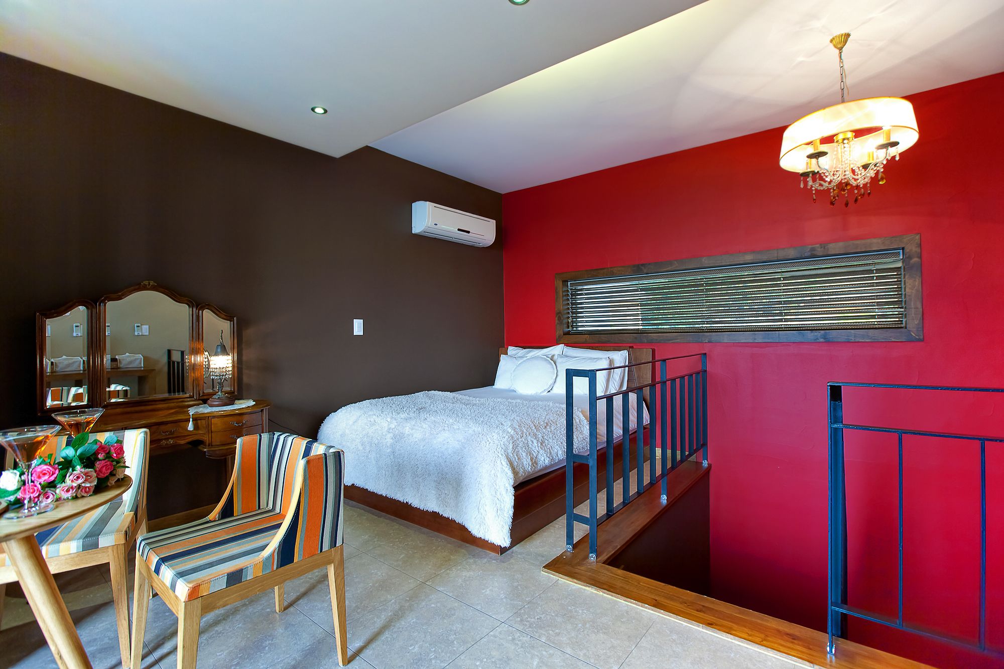 Ein Schlafzimmer mit Bett, Kommode, Stuhl, Tisch mit Blumen, einer Kandelaber und einem Fenster mit Jalousien.