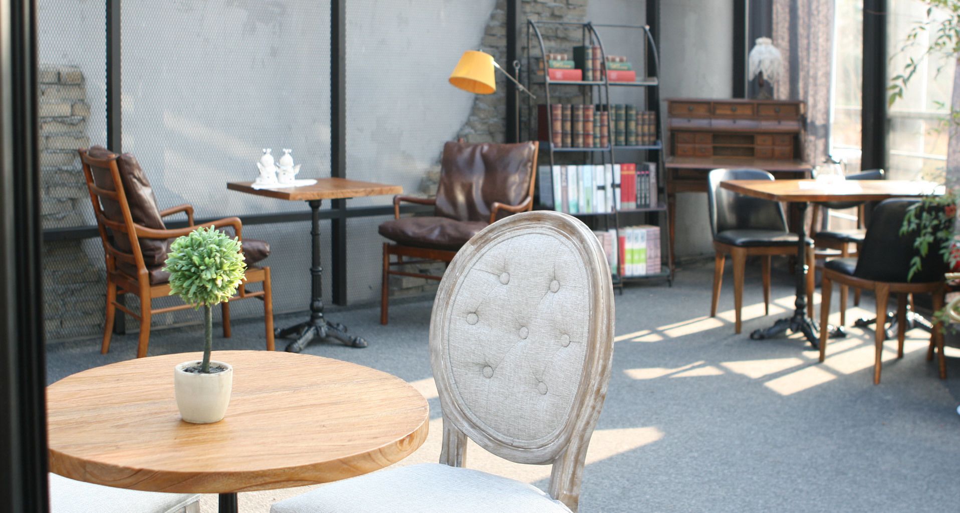 A cozy outdoor seating area with wooden tables and chairs, a small potted plant, and a bookshelf filled with books.