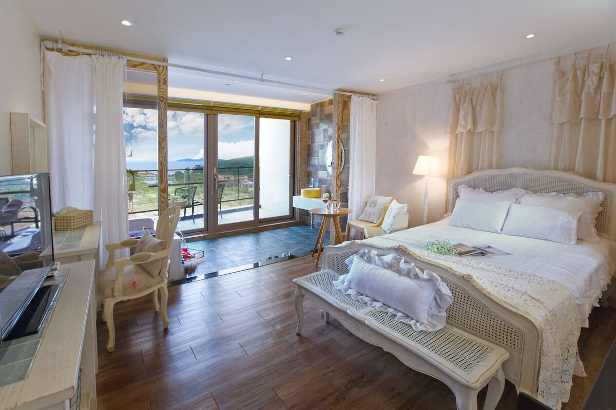 A spacious bedroom with a large bed, a balcony with a view, and a seating area with a table and chairs.