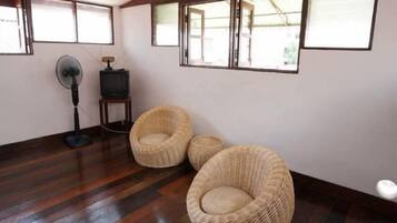 A room with a wooden floor, two wicker chairs, a standing fan, and a television on a small table.
