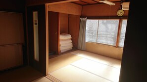 Une pièce de style japonais traditionnel, avec un sol recouvert de tatamis, une pile de futons, une porte coulissante et une fenêtre avec un rideau.