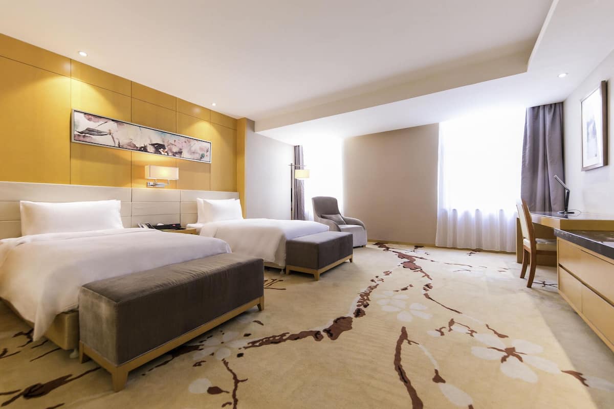 Habitación de hotel con dos camas, un escritorio, una silla, una lámpara y un ventanal grande con cortinas.