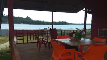 Ein überdachter Außenbereich mit Tisch und orangefarbenen Stühlen, mit Blick auf ein Gewässer und bewaldete Hügel.