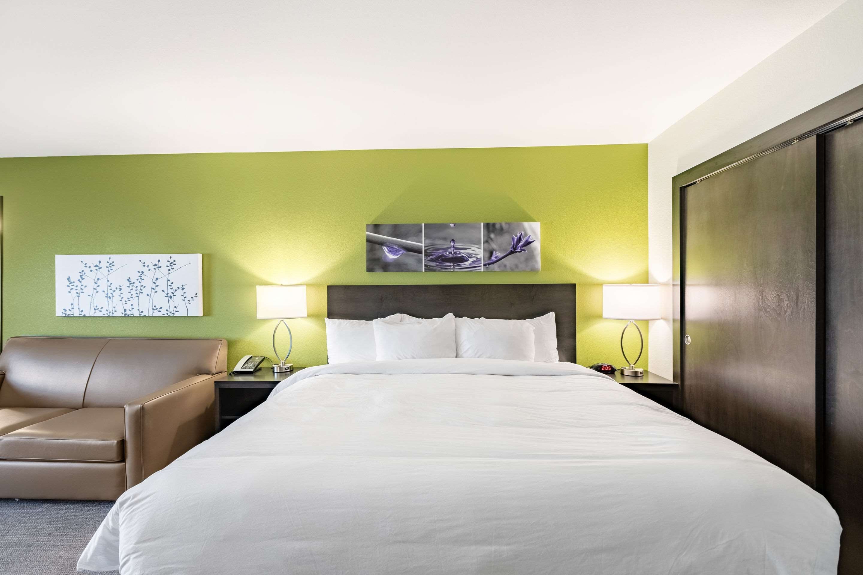 A hotel room with a large bed, a chair, a nightstand with a lamp, and a wall art piece.