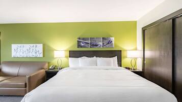 Una habitación de hotel con una cama grande, una silla, una mesita de noche con lámpara y una obra de arte en la pared.
