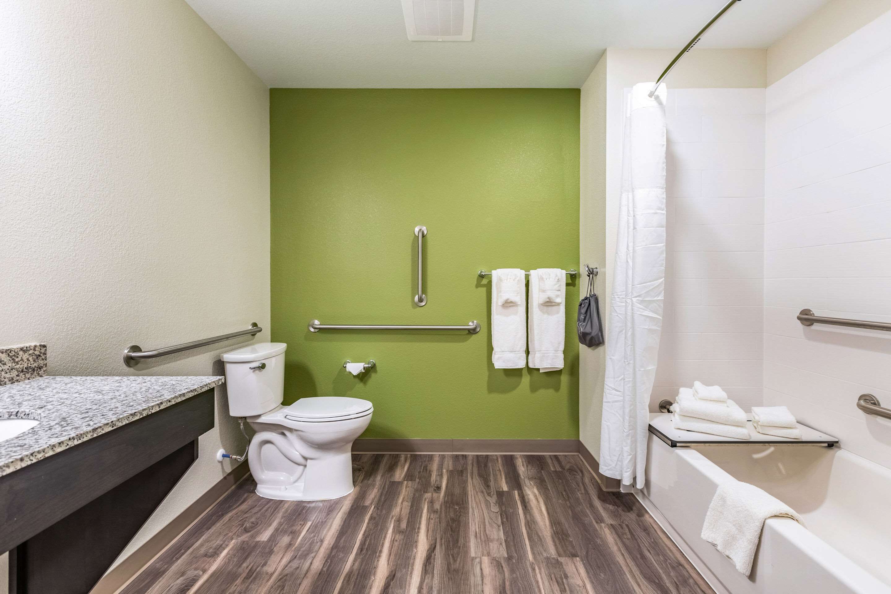A bathroom with a green wall, a white toilet, a sink with a granite countertop, a bathtub with a white shower curtain, and a handrail.