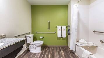 A bathroom with a green wall, a white toilet, a sink with a granite countertop, a bathtub with a white shower curtain, and a handrail.
