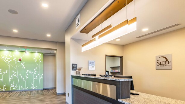 A reception area with a reception desk, a green wall with a floral design, and a sign for Sleep Inn & Suites. - Sleep Inn & Suites Mt. Hope near Auction & Event Center (Mount Hope)