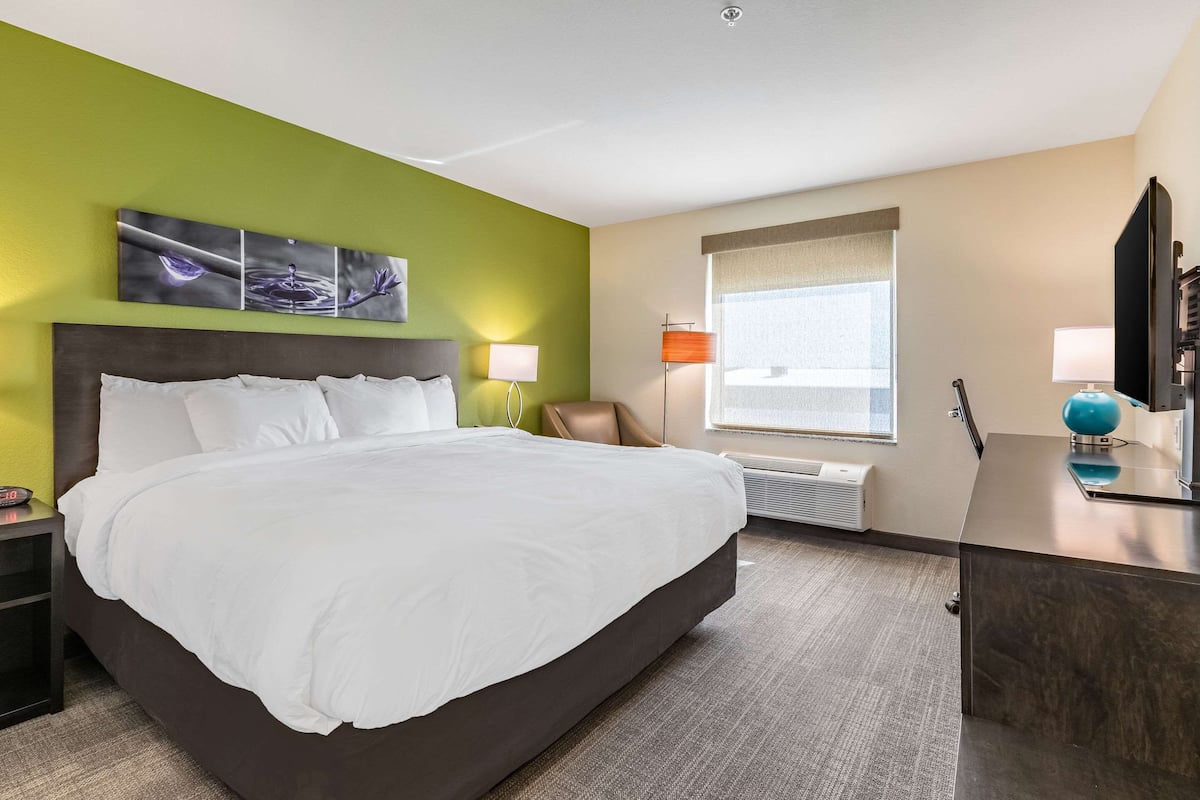 A hotel room with a large bed, a desk with a globe, a chair, a window with blinds, and artwork on the wall.