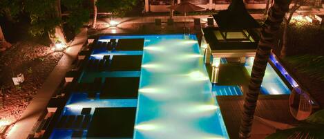 Vue nocturne d’une piscine d’hôtel dotée de plusieurs couloirs de nage, entourée de palmiers et agrémentée d’espaces de détente en plein air.