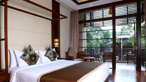 A hotel room with a large bed, a sitting area with a chair and table, and a view of greenery through the window.