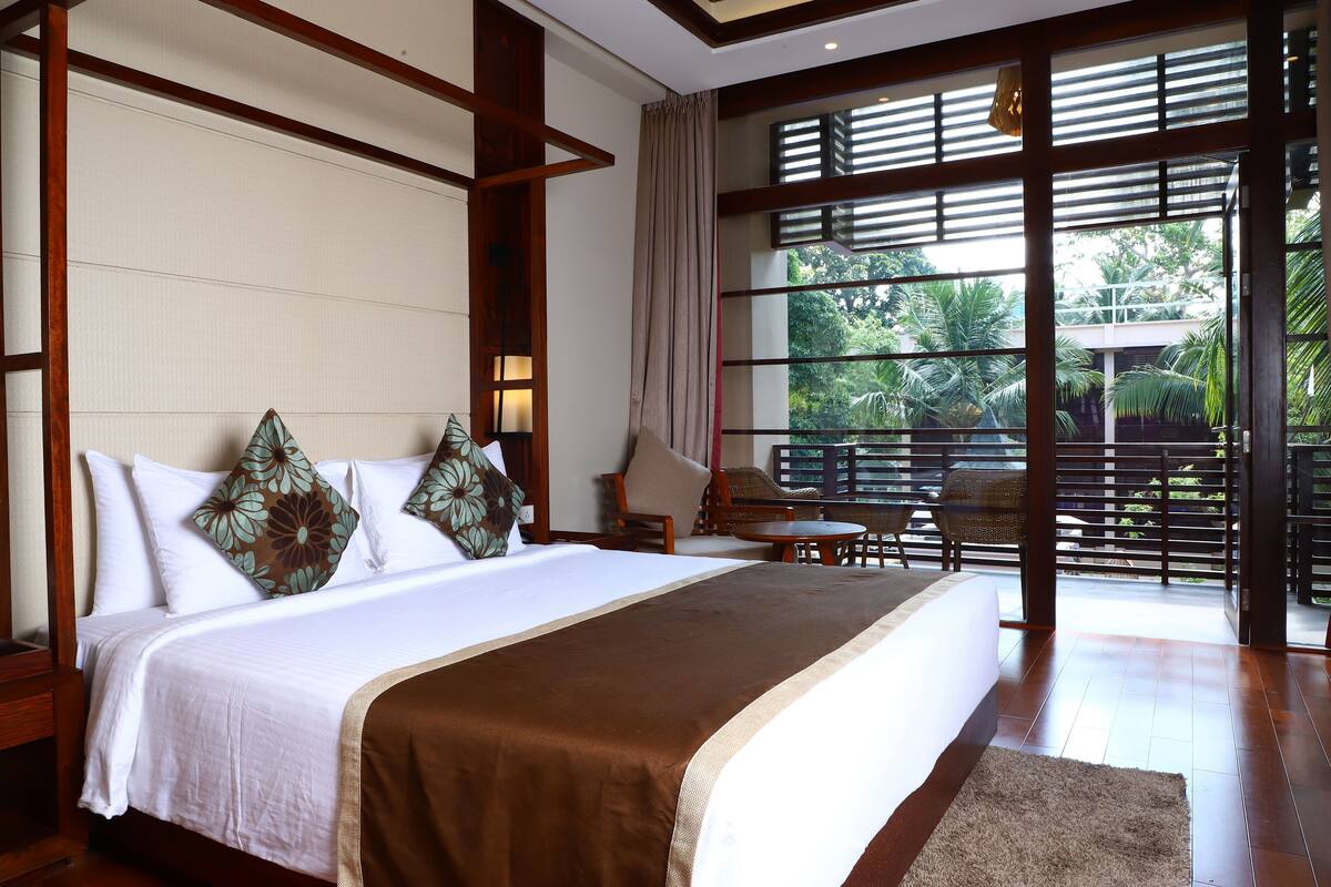 A hotel room with a large bed, a sitting area with a chair and table, and a view of greenery through the window.