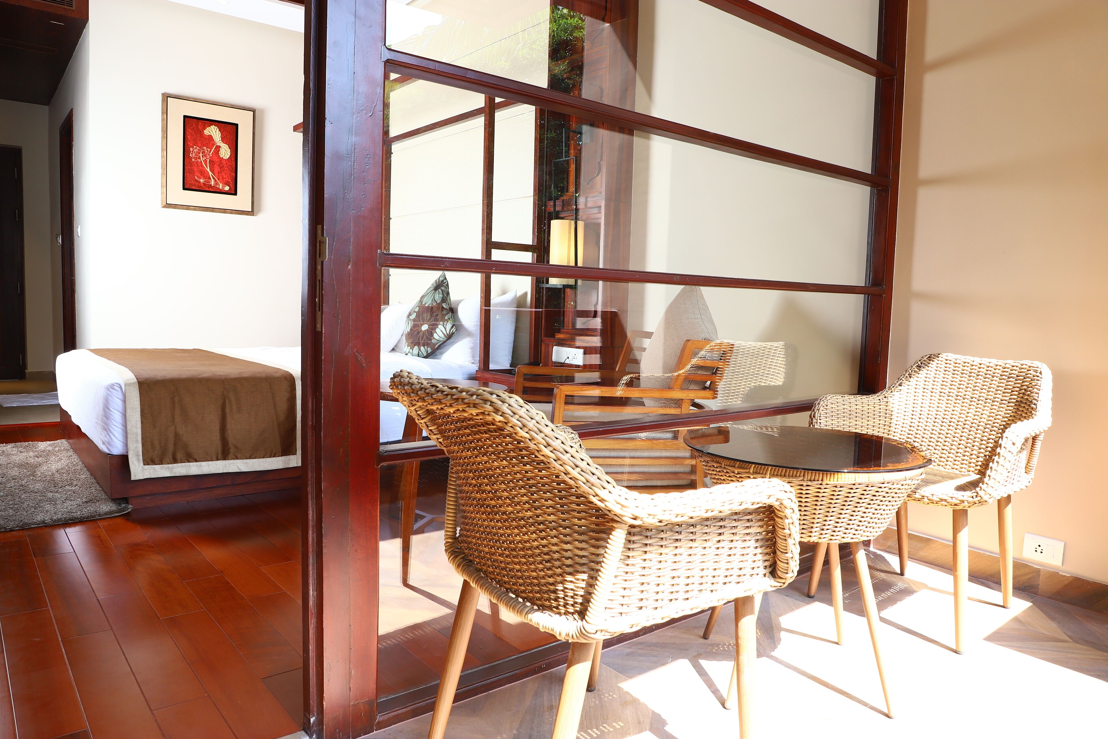 A room with a glass door leading to a balcony, wicker furniture, a bed with a white cover, and a framed picture on the wall.