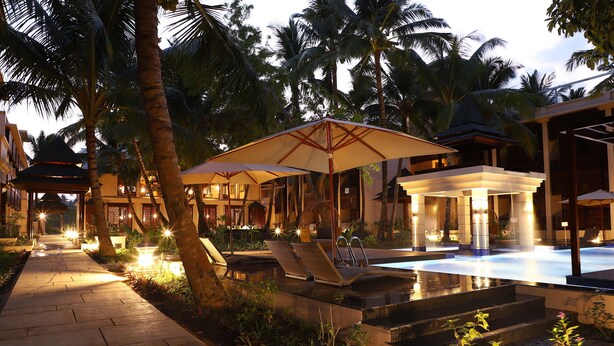 A well-lit outdoor pool area with lounge chairs and umbrellas, surrounded by palm trees and a pathway leading to a building. - Sea Shell Samssara (Port Blair)