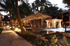 A well-lit outdoor pool area with lounge chairs and umbrellas, surrounded by palm trees and a pathway leading to a building. - Sea Shell Samssara (Port Blair)