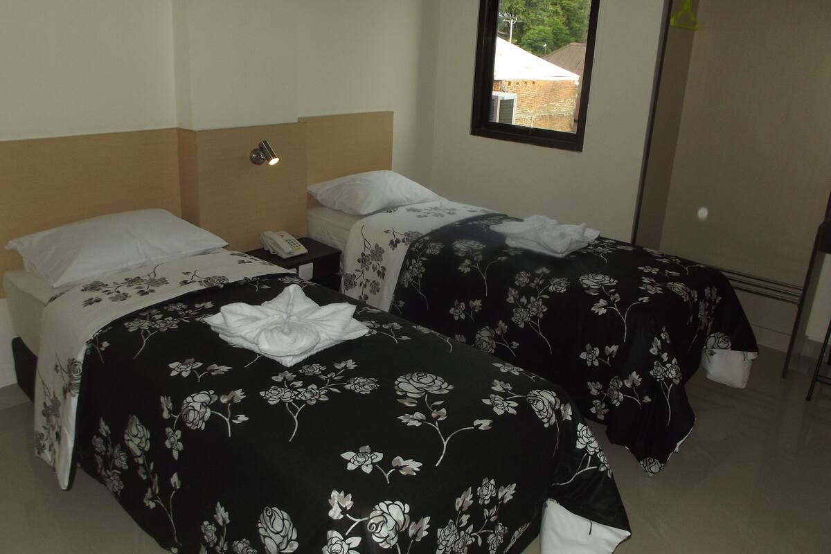 A hotel room with two beds, each with a black floral bedspread and white pillows. There is a small bedside table with a telephone and a wall-mounted light. A window with a view of trees and a building is visible.