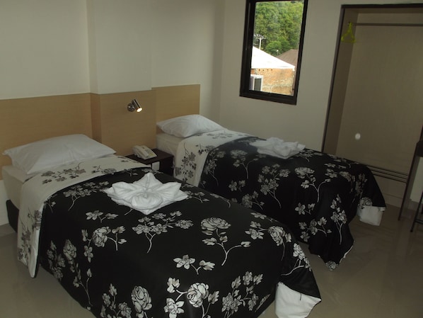 A hotel room with two beds, each with a black floral bedspread and white pillows. There is a small bedside table with a telephone and a wall-mounted light. A window with a view of trees and a building is visible. - Manado Inn Hotel (Manado)