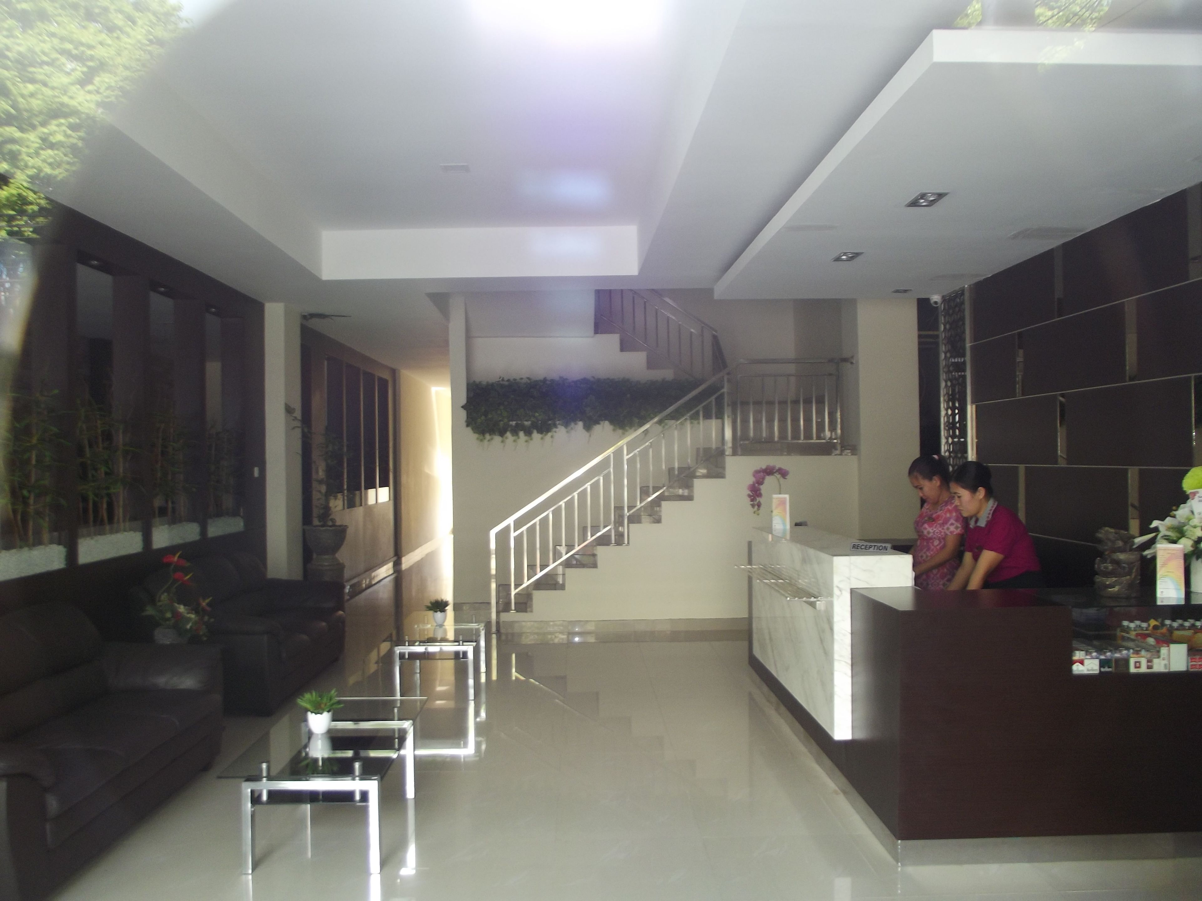 Un área de recepción moderna con mostrador, escalera, sala de estar con sofá y mesa de centro, y un letrero de recepción.