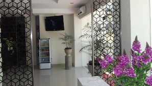 A modern hotel lobby with a geometric patterned partition, a flat-screen TV mounted on the wall, a refrigerator stocked with beverages, and a potted plant. - Manado Inn Hotel (Manado)