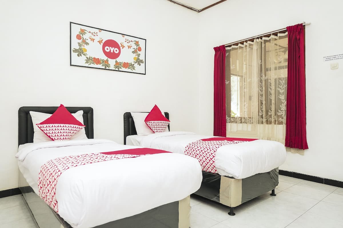 A hotel room with two beds, each with a red and white patterned bedspread and matching pillows. There is a framed floral artwork on the wall above the beds, and a window with red curtains.