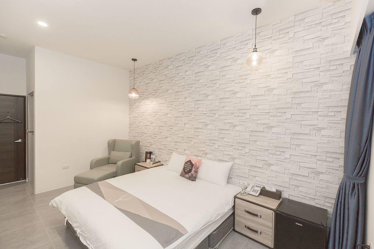 A modern bedroom with a white brick wall, a bed with a white and gray bedspread, a gray armchair, a nightstand with a lamp, and a black Samo appliance.