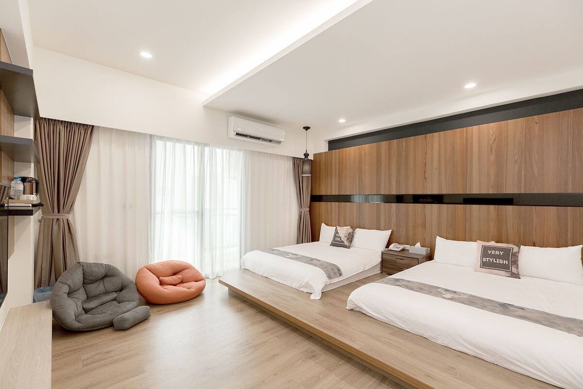 A modern hotel room with two beds, a grey bean bag chair, a wooden headboard, and a large window with curtains.