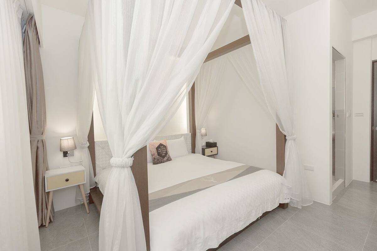 A four-poster canopy bed with a neutral color scheme, a bedside table with a lamp, and a nightstand with a lamp.