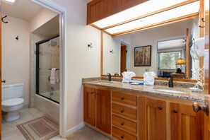 Un baño con cabina de ducha de vidrio, inodoro blanco, armarios de madera, encimera de granito y un espejo grande.