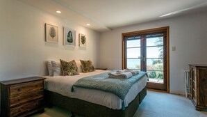 A bedroom with a bed, a wooden dresser, a window with a view, and framed artwork on the wall.