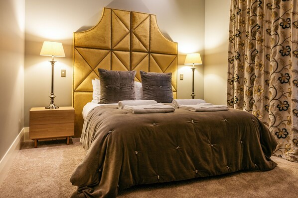 A bedroom with a tufted headboard, two bedside lamps, a wooden nightstand, and a floral curtain. - The Lodge at Mudbrick (Waiheke Island)