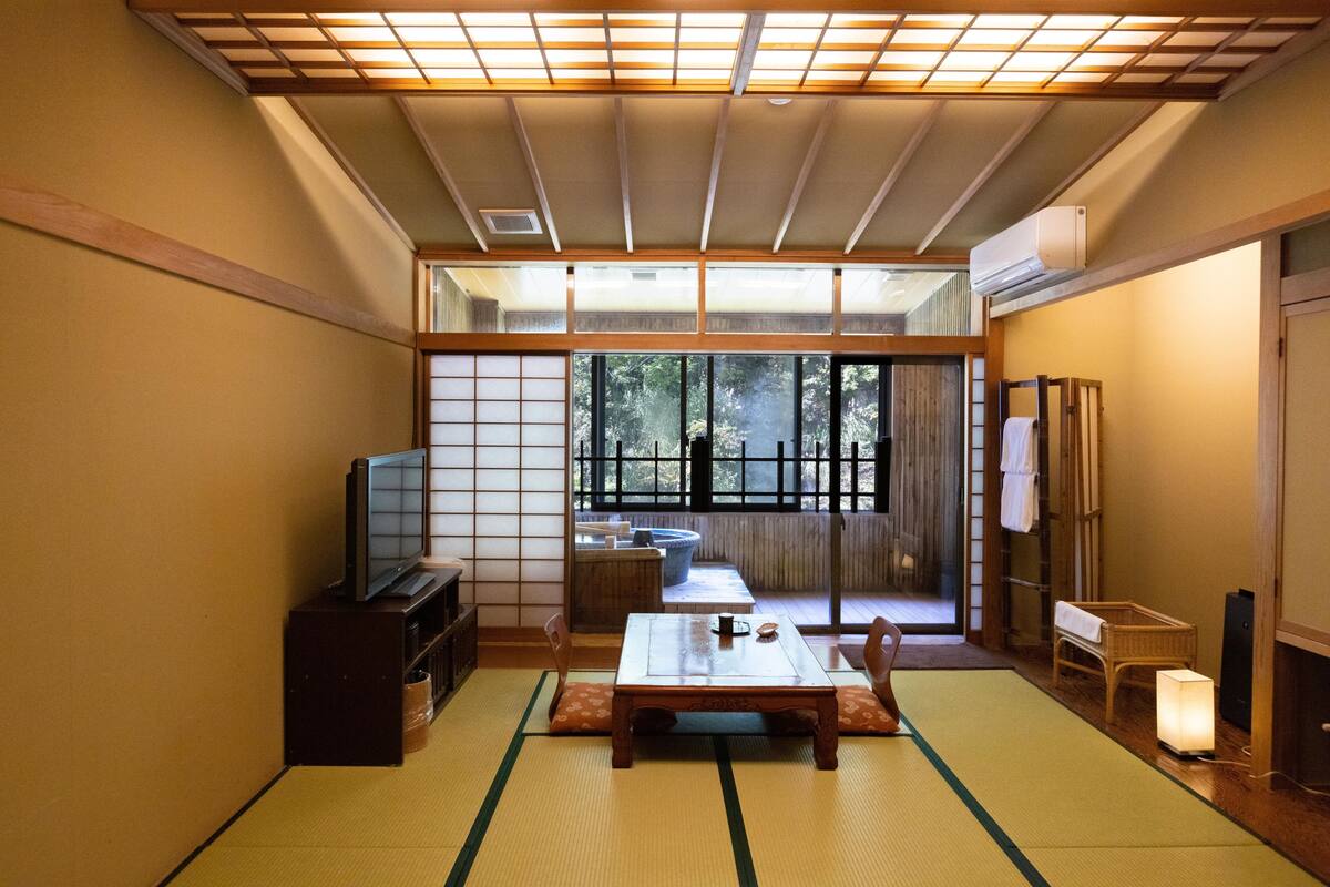 Ett traditionellt japanskt rum med tatamimattor på golvet, ett lågt bord, en tv och en skjutdörr som leder ut till en trädgård.