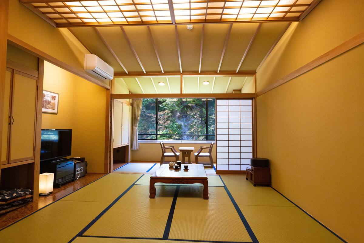 Ett traditionellt japanskt rum med tatamimattor på golvet, ett lågt bord med ett teservis, en tv och en skjutdörr som leder ut till en balkong med utsikt över träd.