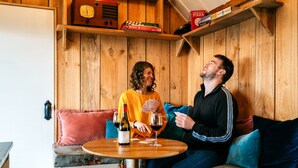 A wooden interior with a round table, a bottle of wine, and playing cards. - The Settlers Hut (Waiheke Island)