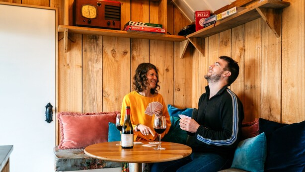 A wooden interior with a round table, a bottle of wine, and playing cards. - The Settlers Hut (Waiheke Island)