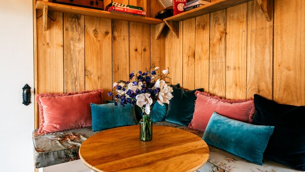 A cozy corner with a wooden bench, a round wooden table, and a vase of flowers. - The Settlers Hut (Waiheke Island)