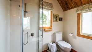 A bathroom with a glass-enclosed shower, a toilet, and a window with a wooden frame. - The Settlers Hut (Waiheke Island)