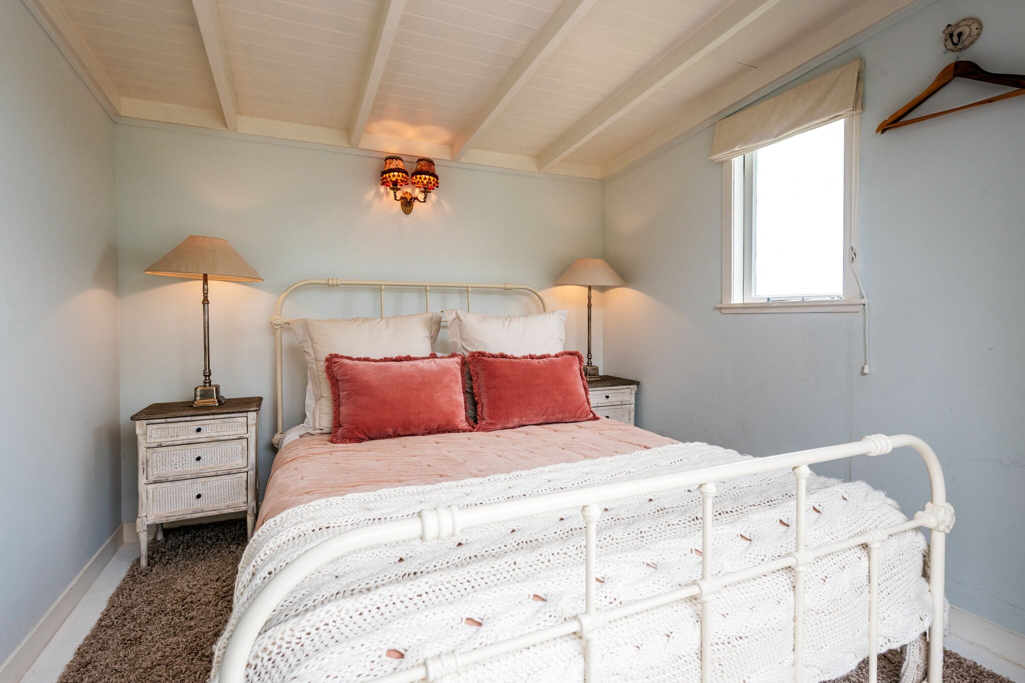 A bedroom with a white metal bed frame, two bedside tables with lamps, a window with a curtain, and a wall-mounted light fixture.