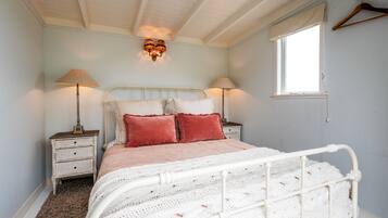 A bedroom with a white metal bed frame, two bedside tables with lamps, a window with a curtain, and a wall-mounted light fixture.