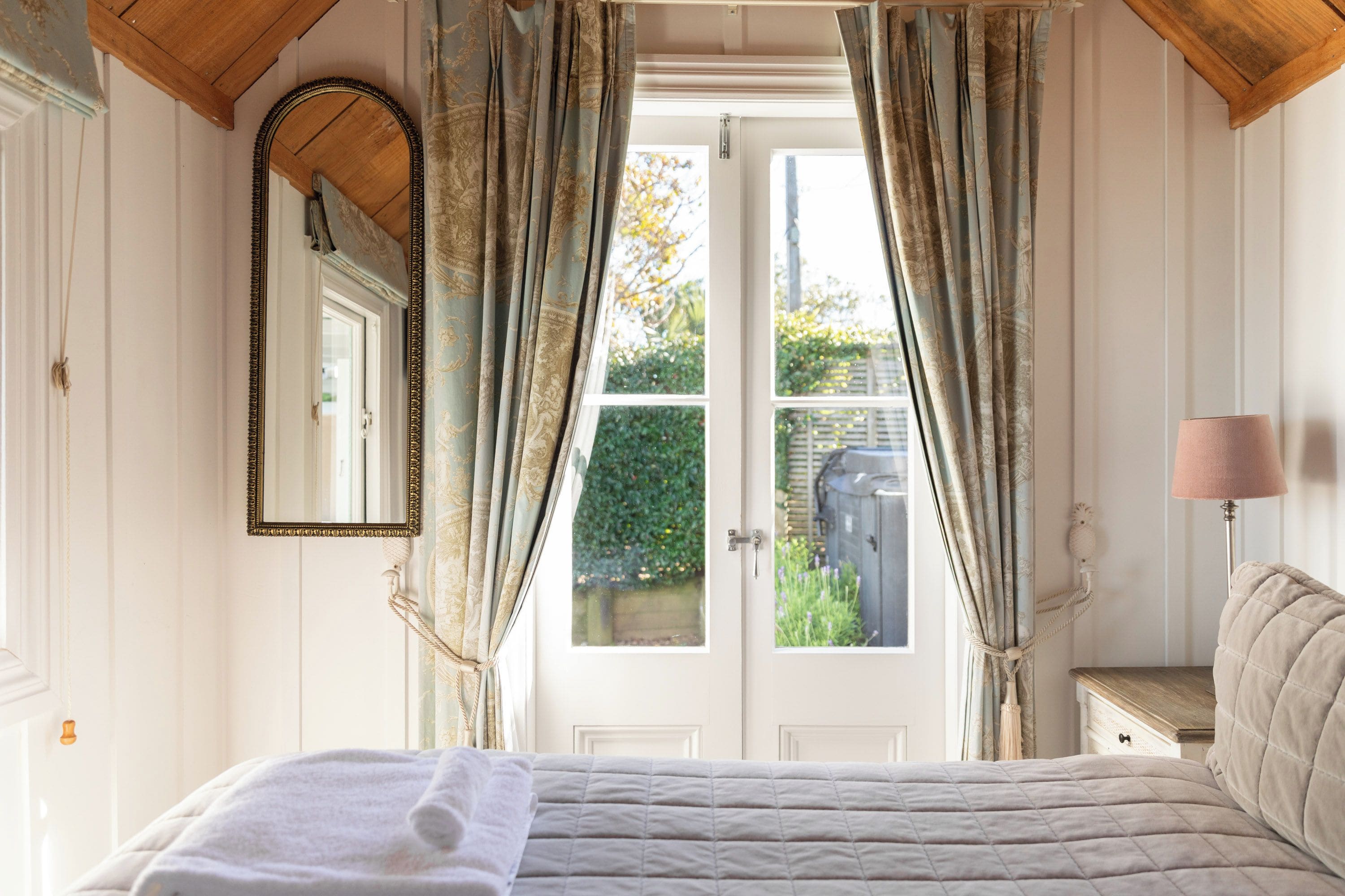 A bedroom with a bed, a mirror, curtains, and a view of the outside.