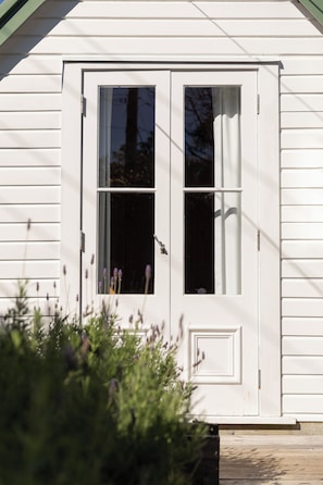 A white wooden door with a window, a small porch, and a plant in the foreground. - The Settlers Hut (Waiheke Island)