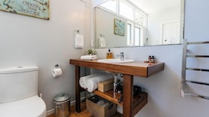 A bathroom with a wooden vanity, a toilet, a trash can, and a towel rack. - The Vintage Cottage (Waiheke Island)