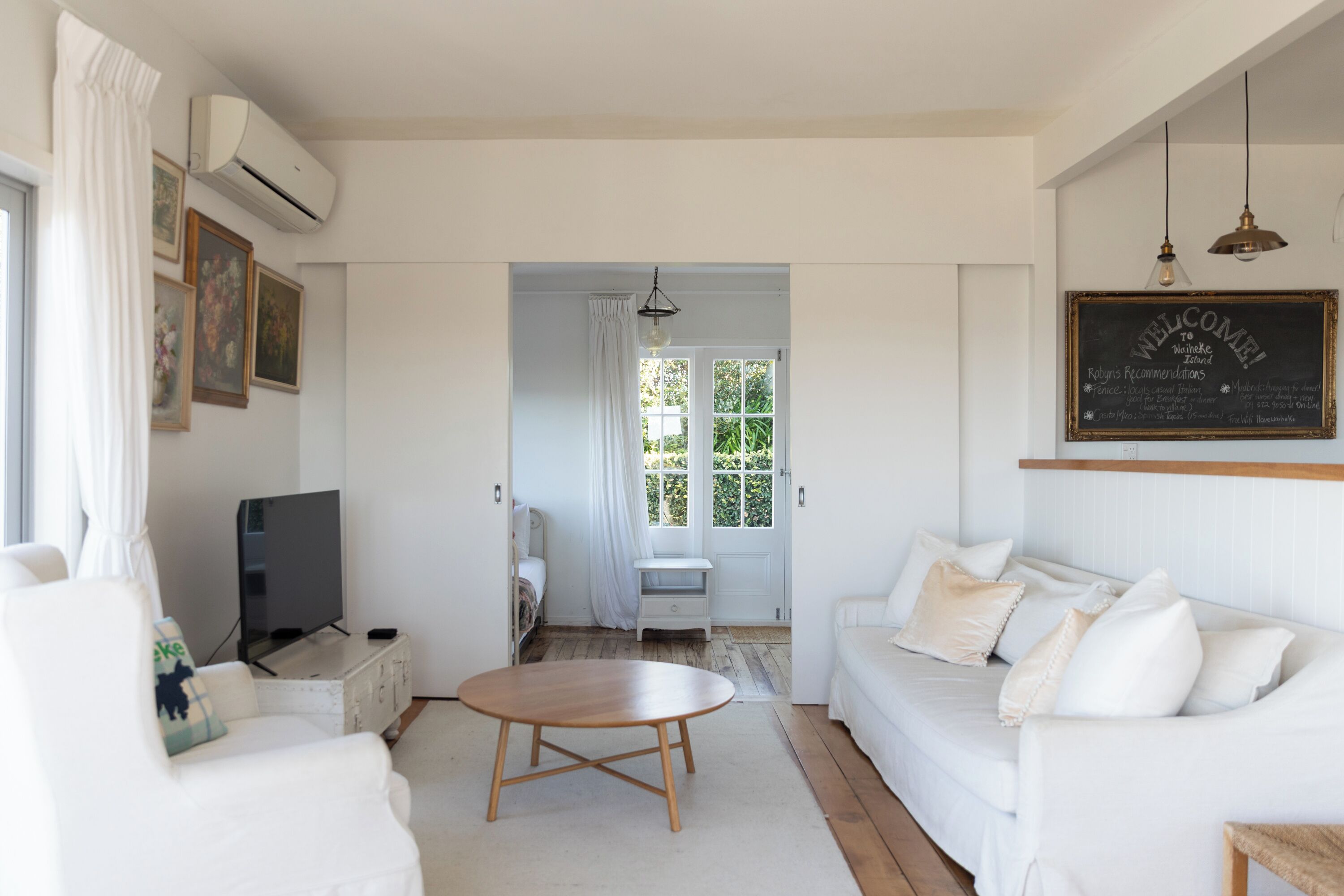 A spacious living room with a white sofa, a wooden coffee table, and a television. There is a chalkboard with 'WELCOME TO Wainkae Island' written on it.