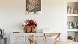 A dining area with a wooden table and chairs, a white cabinet with a floral painting above it, and a bookshelf with books and decorative items. - The Vintage Cottage (Waiheke Island)