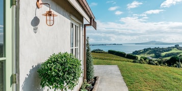 Uma casa com uma janela grande, luminária fixada na parede, um pequeno jardim e vista para um lago e colinas.