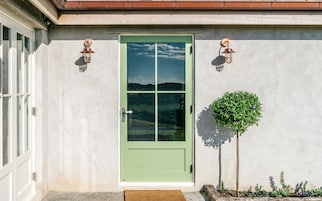 Uma porta verde com uma vista clara do céu e da paisagem através dos painéis de vidro, com duas luminárias fixadas na parede e uma pequena árvore ao lado da porta.