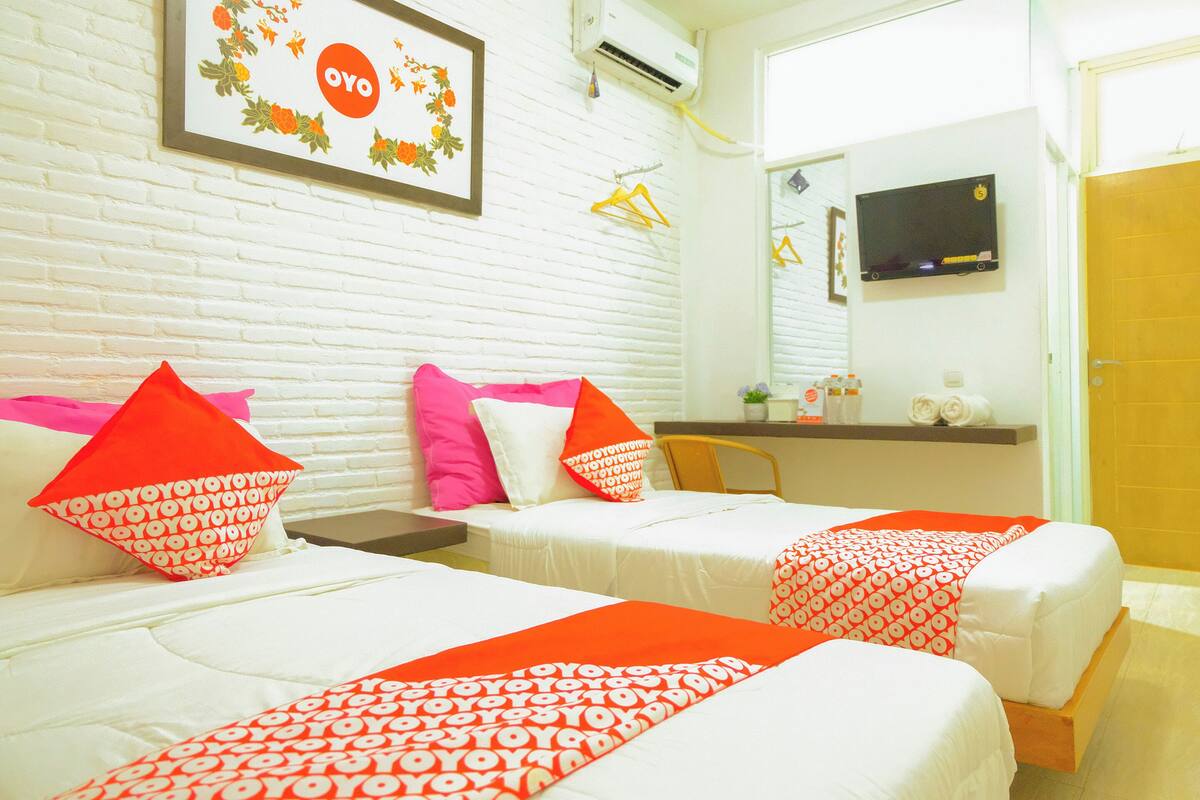 A hotel room with two beds, each with white bedding and orange pillows. There is a framed picture on the wall, a TV mounted above a wooden dresser, and a mirror on the wall.