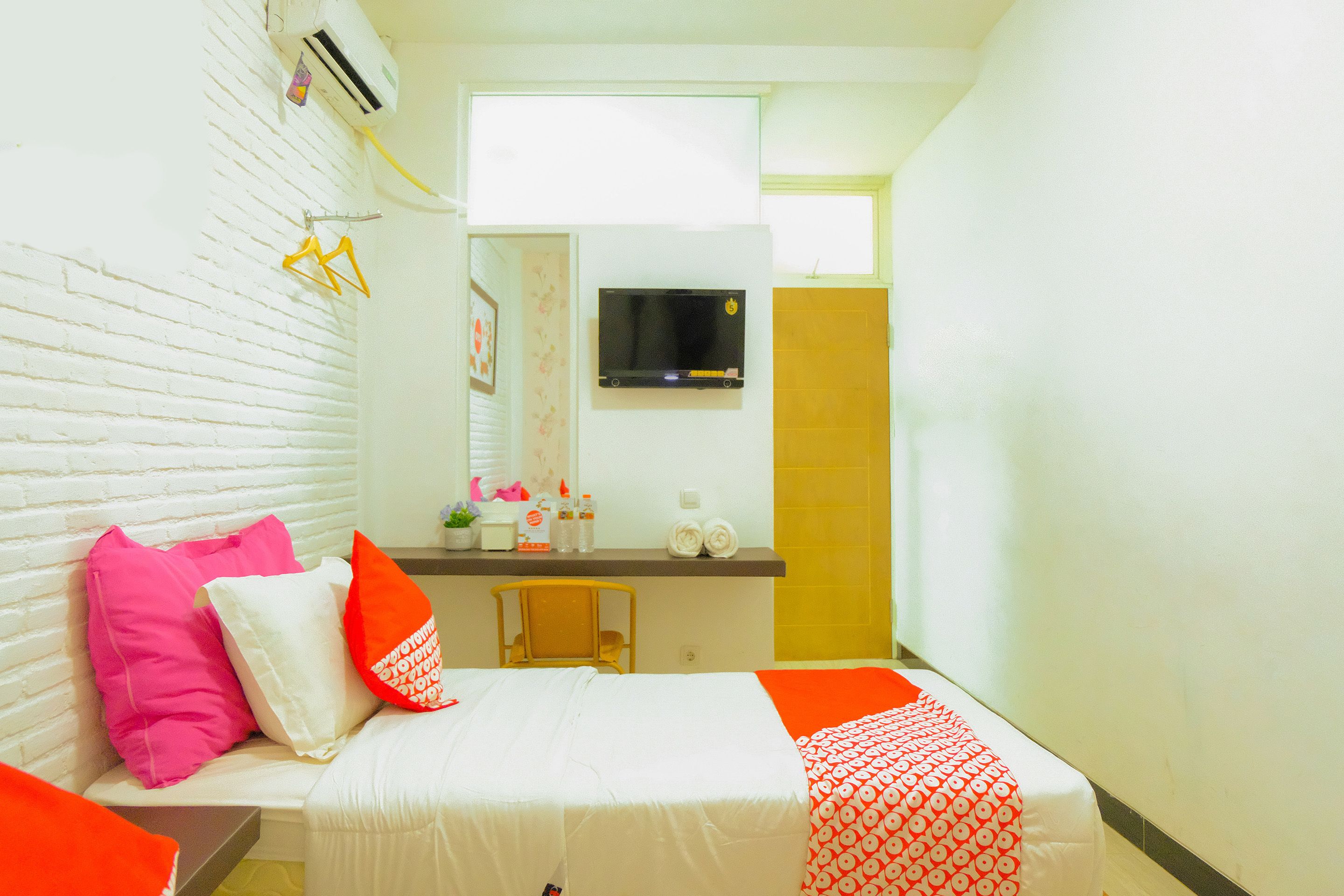 A small, brightly lit room with a bed, a small table with toiletries, a mirror, and a TV mounted on the wall.