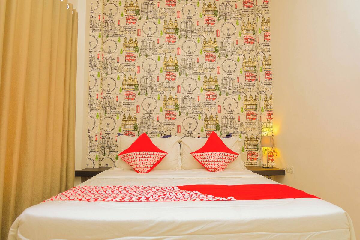 A hotel room with a patterned wallpaper, a bed with red and white bedding, and two red pillows.
